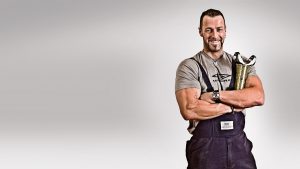 Portrait of muscular man holding metal product against a grey background, wearing a grey Umbra t-shirt and dark blue dungarees, smiling at camera.