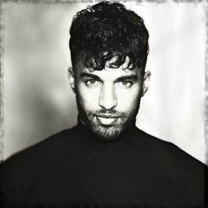 Black and white head shot of Thomas King male model wearing black crew neck sweater. model has dark short curly hair and is looking intensely into camera. The background is mainly white dappled with grey on the left side. The edge of the image is framed with a faded broken border, ala polaroid.