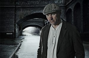 Portrait of man in peaked cap and Edwardian style clothes standing in front of a Victorian brick viaduct. The background is dark, wet and deserted, a working class Edwardian/Victorian Street scene.