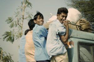 Four boys on the way to work, hanging off the back of a light green Land Rover smiling at the camera.
