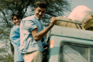 Image of two indian boys in blue shirts hanging off the back of a light green land Rover one of them flexing his muscles. On the way to work
