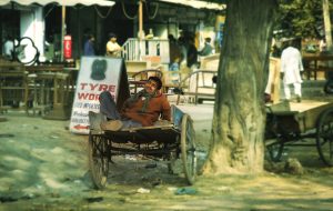 An Indian man asleep in cart wearing brown top and blue trousers and green scarf, Delhi, India