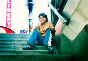 Young girl sitting on steps of subway wearing blue trousers and orange top, sporting self drawn tattoos with her thumb up